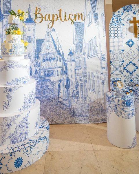 Montaje de bautizo en azul y blanco: fondo de calles ilustradas, torta decorativa de varios niveles con flores y pedestal con azulejos