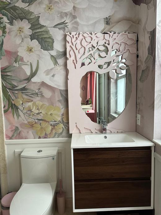 Baño con papel tapiz de flores y un panel rosado con árbol calado alrededor de un espejo ovalado, sobre un lavabo blanco